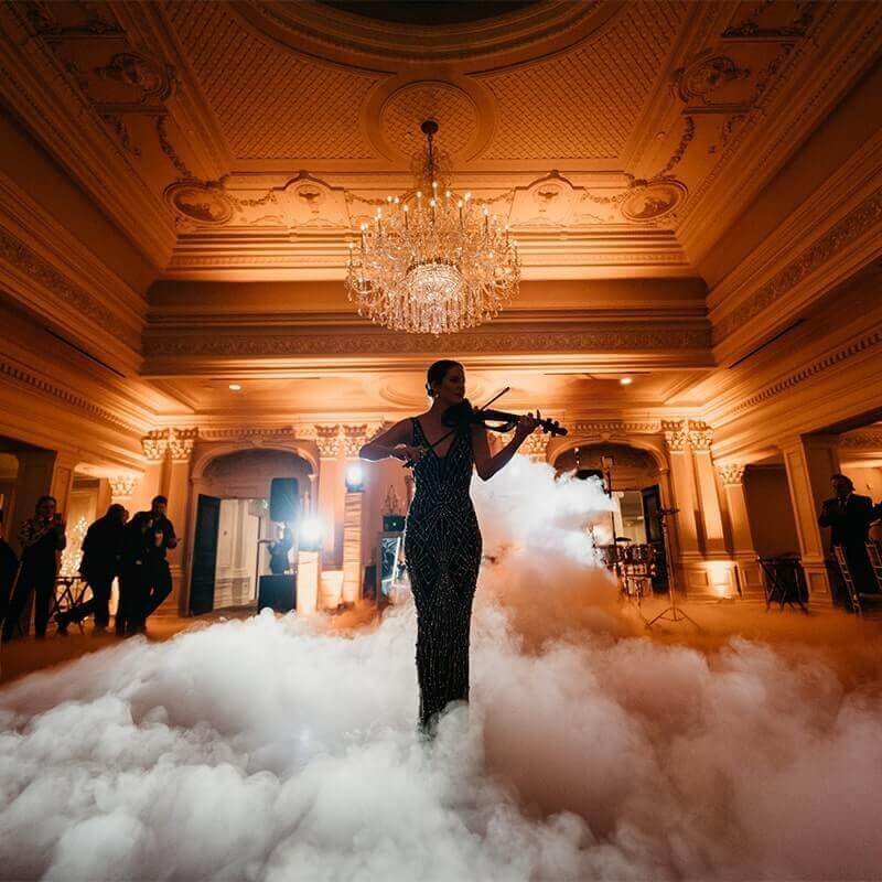 Event-Services A woman in a formal dress plays violin in an ornate, chandelier-lit hall filled with low-lying fog, while people watch in the background.
