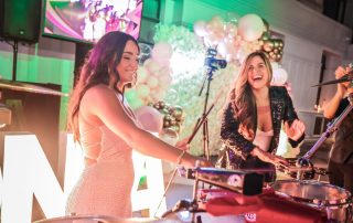 Two women smiling and playing drums at a brightly lit event with balloons, a camera, and a TV screen in the background.
