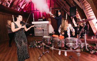 A woman plays the violin and another plays percussion at an indoor event, with smoke effects and people in the background near a DJ booth.