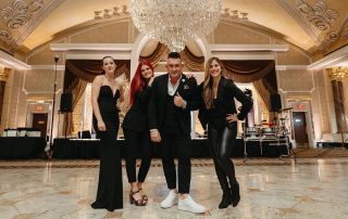 Four people dressed in formal and semi-formal attire pose together in an elegant, chandelier-lit ballroom with ornate architecture.