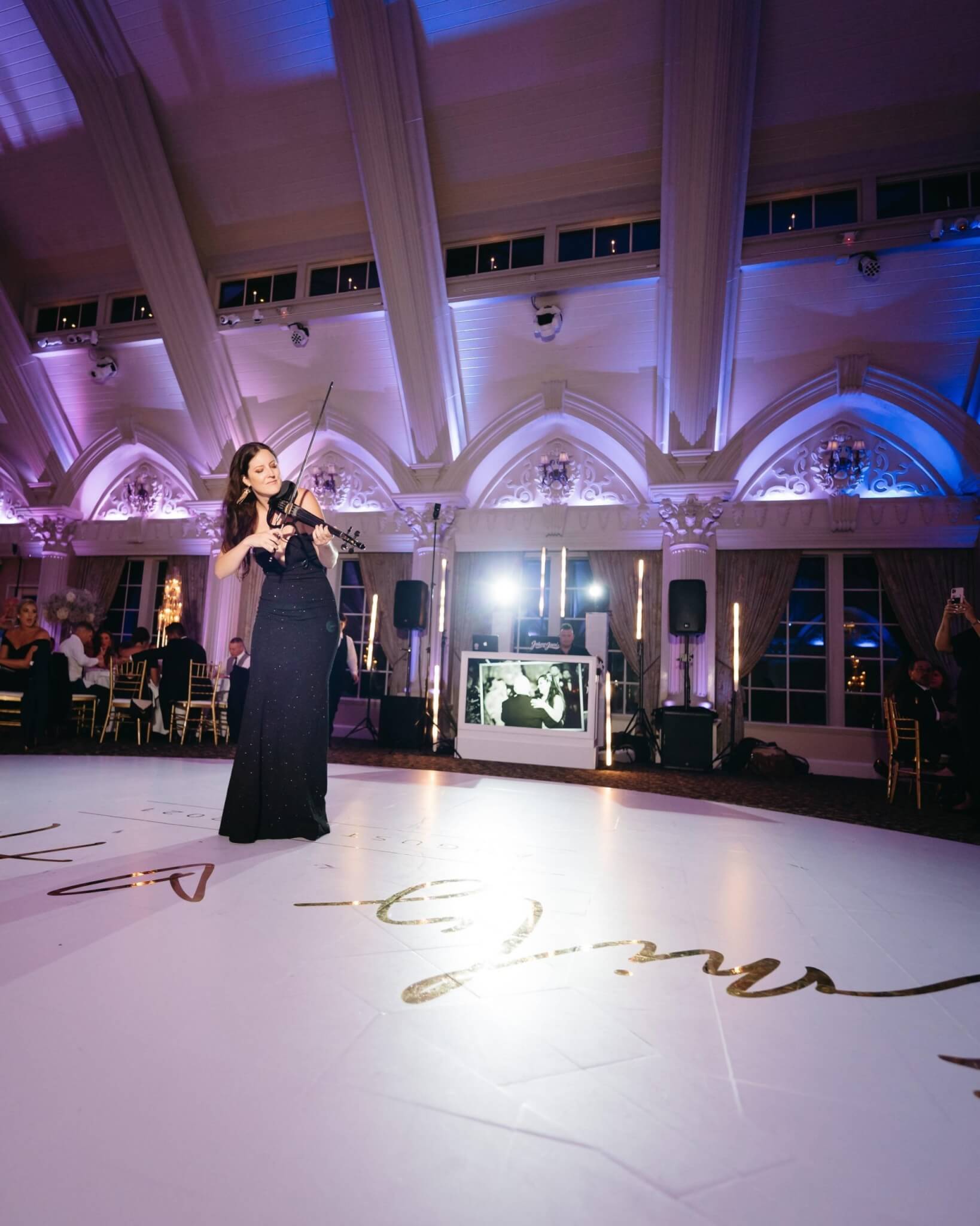 A woman in a black dress plays the violin on a large decorated floor in an elegant, high-ceilinged event hall with purple lighting.