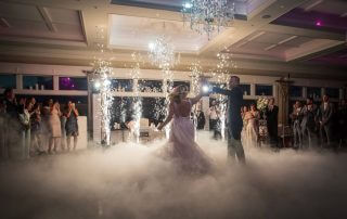 A bride and groom dance on a fog-covered floor with sparklers and guests watching in a decorated indoor venue.