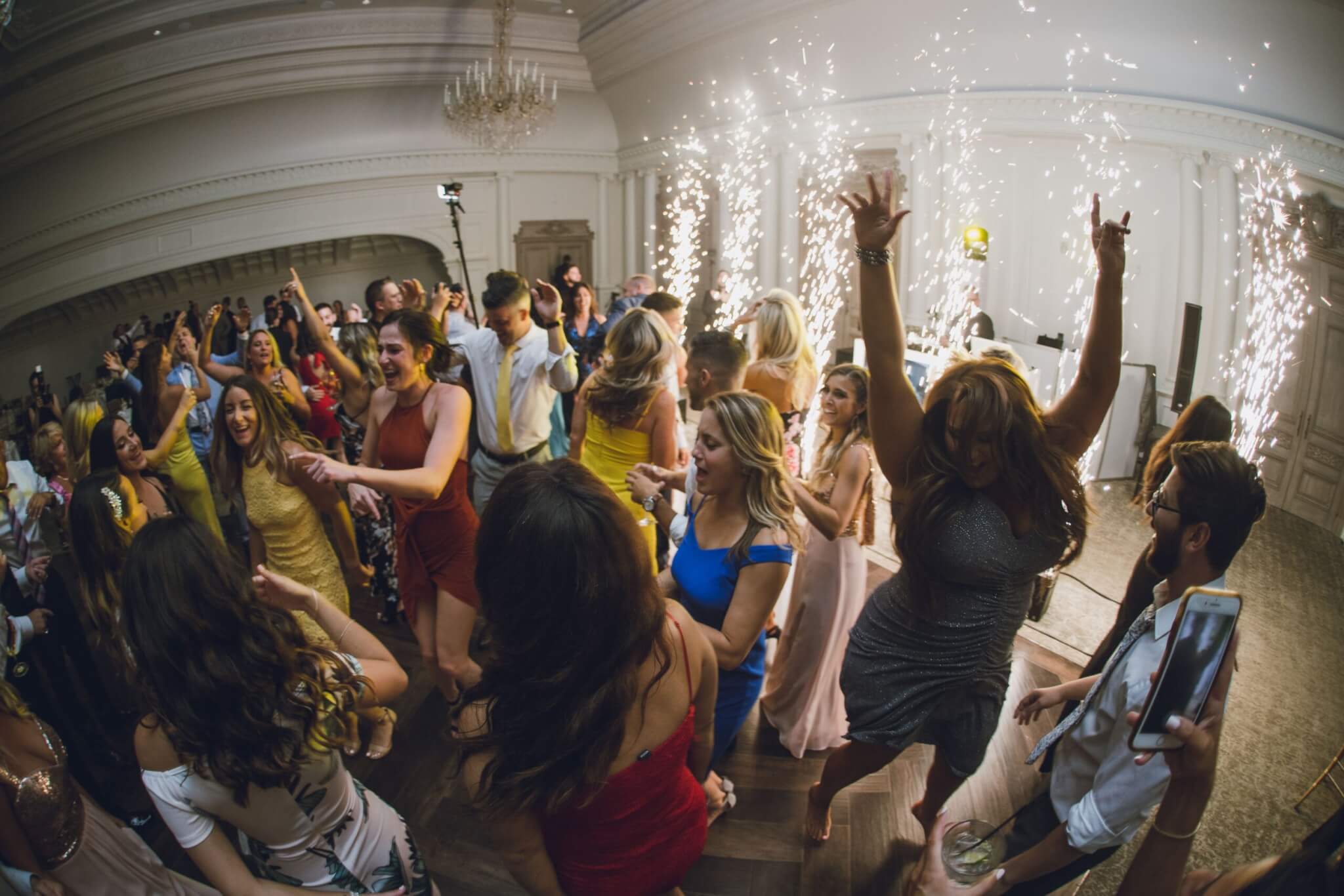 A large group of people dance and celebrate indoors, surrounded by sparklers and festive lighting in a bright, elegant room.