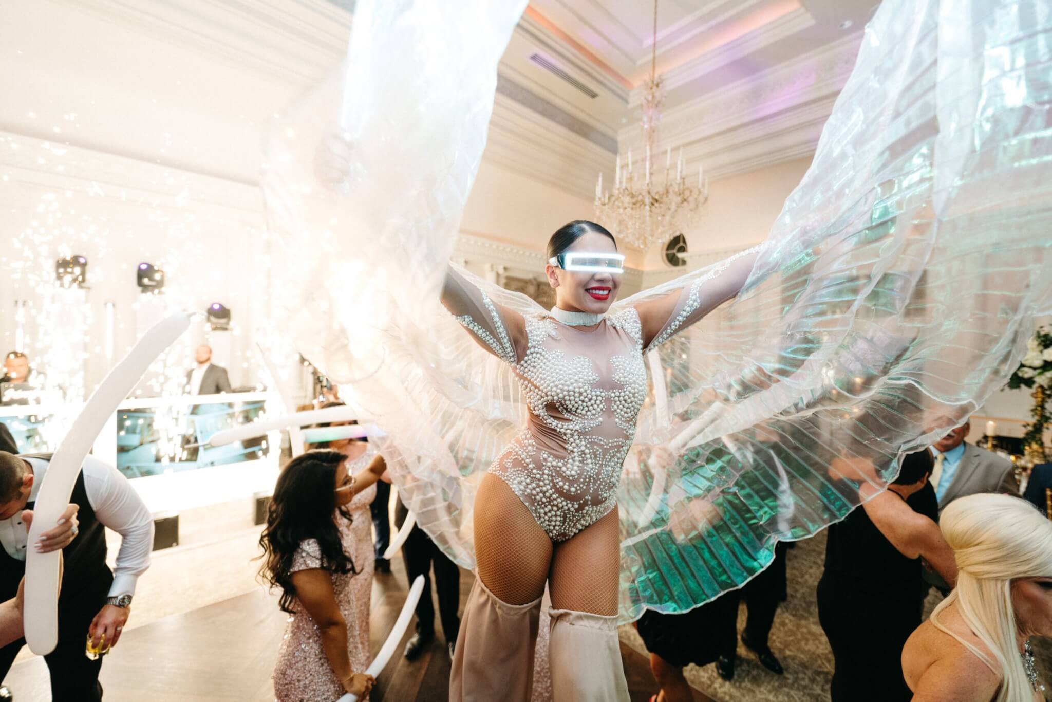 Performer in a glittery bodysuit with large translucent wings entertains guests at an indoor event; people are dancing and lights are flashing.