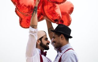 Two men embrace outdoors, holding several large red heart-shaped balloons above their heads.