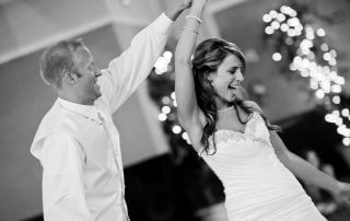 A bride and groom dance together in formal attire, smiling, with string lights blurred in the background.