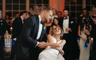 A groom in a black tuxedo dips his smiling bride in a white dress during their wedding reception, surrounded by guests in formal attire.