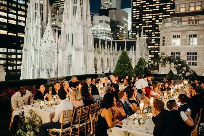 A group of people seated at long, candlelit tables on a rooftop terrace at night, with city buildings and a large cathedral visible in the background.
