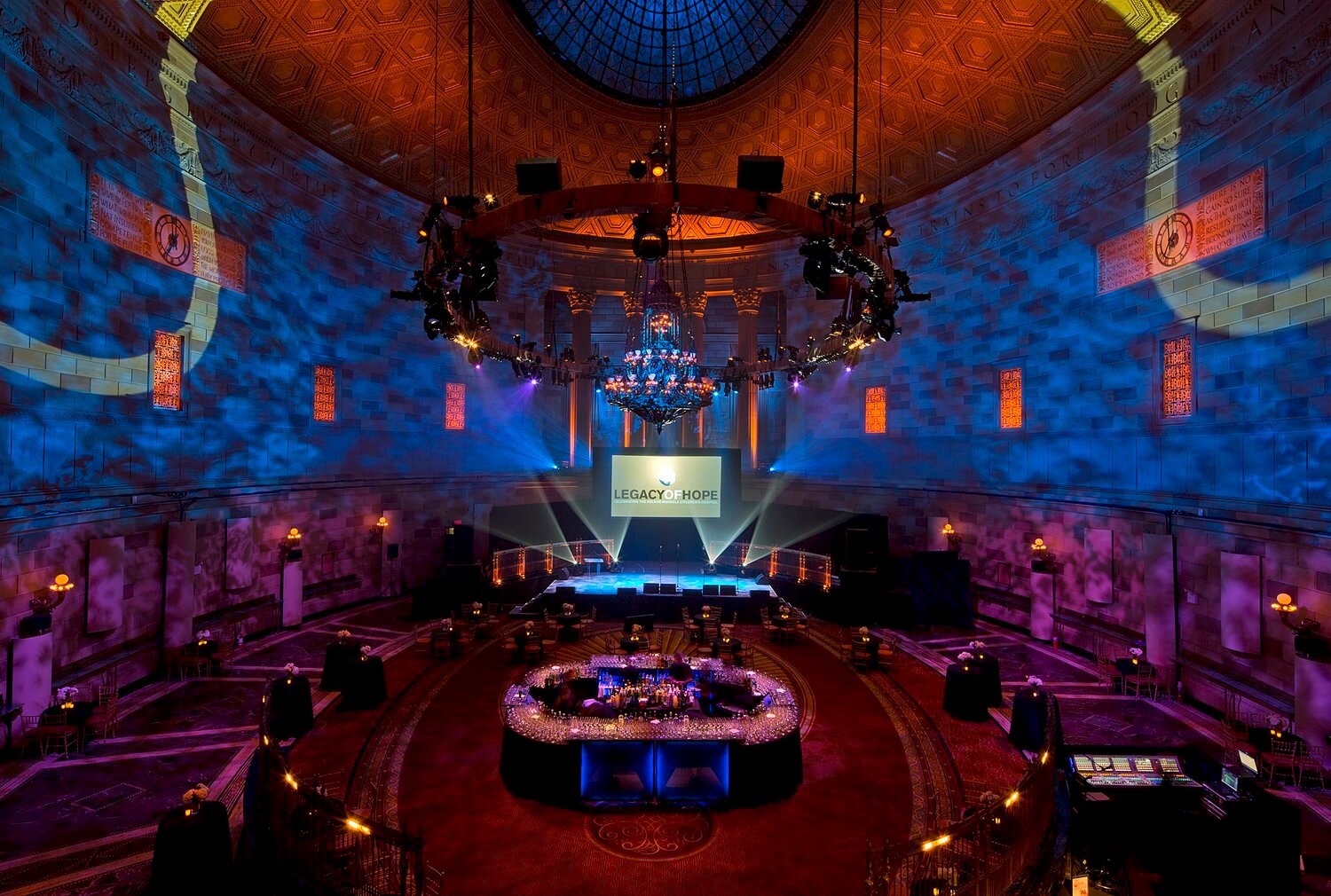 Large, ornate event hall with a domed ceiling, dramatic blue lighting, central chandelier, round bar, stage with a screen displaying “Legacy of Hope,” and empty tables.