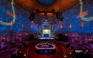 Lavishly decorated ballroom set up for an event, featuring a central chandelier, stage with a screen reading "Legacy of Hope," and round dining tables under dramatic blue lighting.