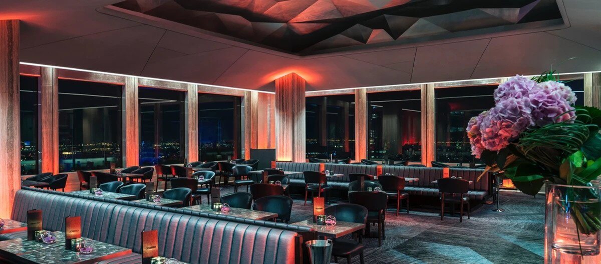 Modern rooftop restaurant with floor-to-ceiling windows, ambient lighting, black leather seating, and city views at night; a vase of pink flowers is in the foreground.