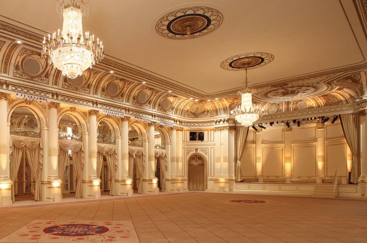Large, ornate ballroom with chandeliers, high decorated ceilings, arched windows, and patterned carpet, all in cream and gold tones.