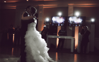 A bride and groom share a first dance in a dimly lit reception hall, with guests and illuminated screens in the background.