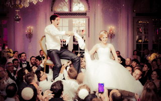 A bride and groom are lifted on chairs by guests during a wedding celebration inside a decorated hall.