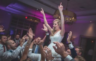 A bride in a white dress is lifted in the air by a group of cheering guests at a lively indoor wedding reception.