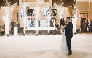 A bride and groom dance together on a fog-covered floor, surrounded by indoor fireworks, at a decorated wedding reception venue.