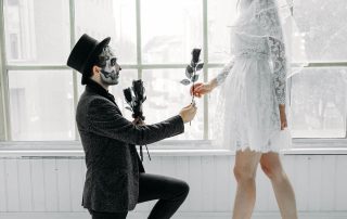 A person in a suit and top hat kneels and offers a black rose to another person in a white lace dress and veil, both with face paint, in front of a large window.