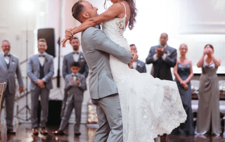 A groom lifts the bride during their first dance at a wedding reception, with guests standing and watching in the background.