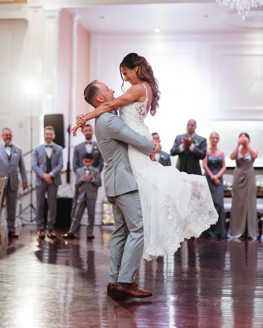 A groom in a gray suit lifts the bride in a white dress on a dance floor, with guests in formal attire watching and applauding in the background.
