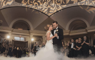 A bride and groom share their first dance on a cloud-covered floor in an ornate ballroom, surrounded by elegantly dressed guests.