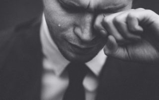 A man in a suit wipes his eyes as a tear rolls down his cheek; the image is black and white with a close-up focus on his face and hand.