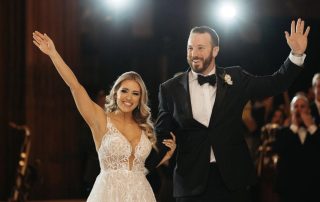 A bride in a white lace gown and a groom in a black tuxedo stand arm in arm, smiling and waving at an indoor event with bright lights in the background.