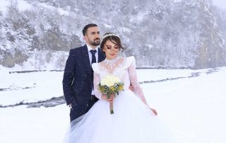 A bride in a white dress and groom in a suit stand together in a snowy outdoor landscape with mountains in the background.