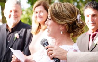 A woman in a white dress speaks into a microphone while holding a paper, with three people smiling in the background at an outdoor event.