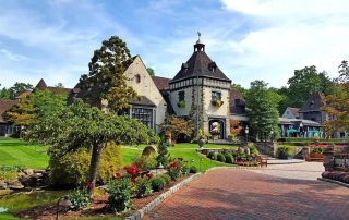A large stone building with a peaked roof and clocktower sits amid manicured lawns, flower beds, and brick pathways on a sunny day.