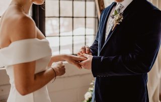 A groom places a ring on the bride’s finger during a wedding ceremony by a window.