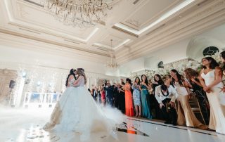 Two brides kiss on a decorated dance floor as guests watch and take photos in an elegant, chandelier-lit wedding venue.