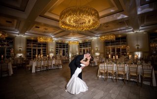 A bride and groom share a kiss while dancing in the center of an elegantly decorated, empty banquet hall with chandelier lighting.