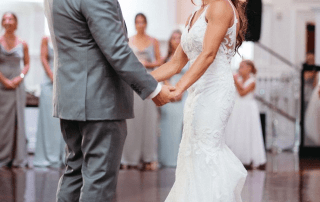 A bride in a white gown and an older man in a gray suit hold hands and smile while dancing at a wedding reception, with bridesmaids watching in the background.