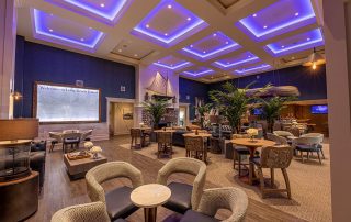 Spacious lounge with modern decor, blue LED ceiling lights, wooden tables, cushioned chairs, indoor plants, and nautical-themed accents.