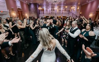 A bride in a white dress dances with a man at the center of a crowded wedding reception, with guests clapping and celebrating around them in a decorated ballroom.