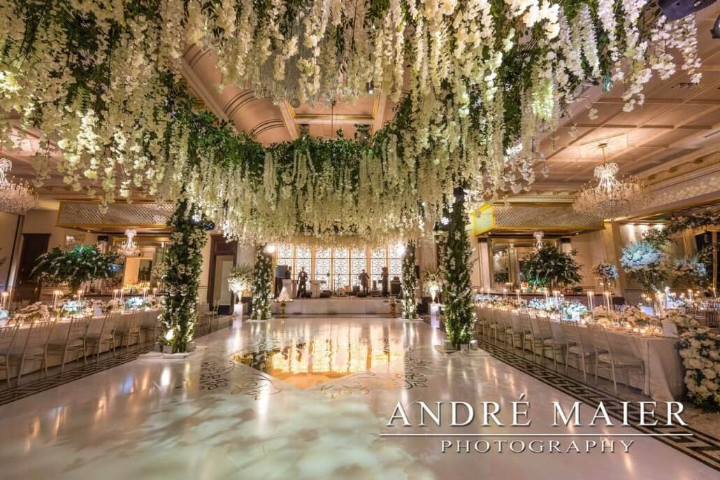 Elegant ballroom decorated for a wedding with cascading white flowers, long banquet tables, and lavish lighting; photo by André Maier Photography.