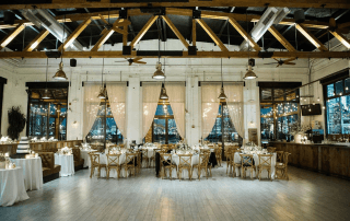Spacious event hall with exposed wooden beams, round tables set for dinner, white drapes, string lights, and large windows overlooking a cityscape.