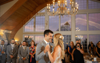 A bride and groom share their first dance at a wedding reception in a hall with large windows and a chandelier, surrounded by standing guests.
