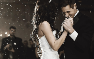 A bride and groom share a first dance at their wedding, holding hands and smiling, with guests and soft lights in the background.