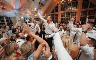 A large group of people dressed in formal attire dance and raise their arms at an indoor event, as one man stands in the center holding up a phone.