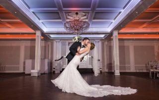 A groom in a suit dips and kisses a bride in a white gown on the dance floor of an elegant, chandelier-lit ballroom.