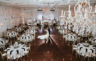 A bride and groom stand together in the center of a large, elegant banquet hall with round, set tables and chandeliers overhead.