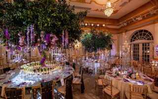 Elegant banquet hall with round tables, gold chairs, tall glass centerpieces, and lush hanging floral arrangements in purple and pink tones under a chandelier.