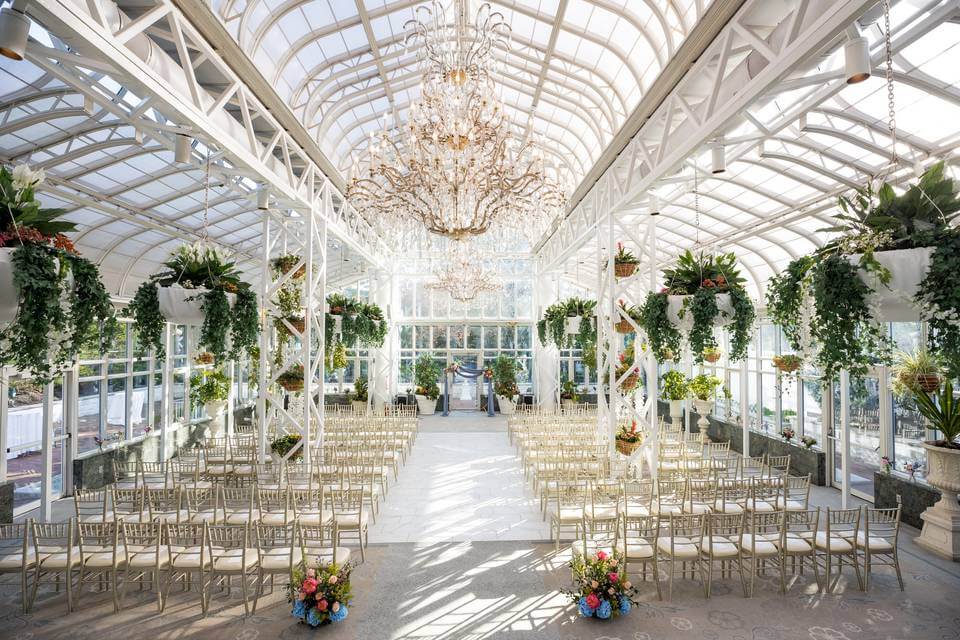 A bright indoor wedding venue with rows of gold chairs, hanging greenery, large chandeliers, and floral arrangements under a glass ceiling.