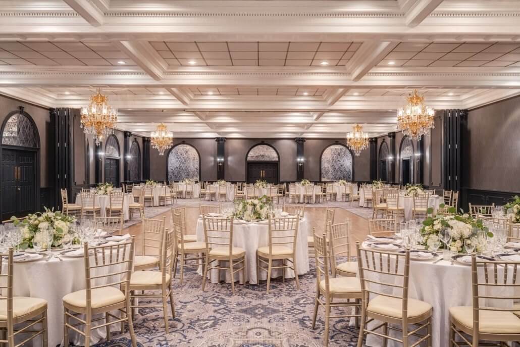 Elegant banquet hall with round tables set for an event, gold chiavari chairs, floral centerpieces, chandeliers, and a patterned carpet.