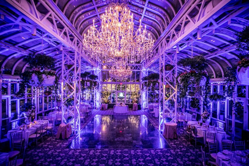 A grand, glass-ceilinged hall decorated with chandeliers, purple lighting, plants, and round dining tables arranged around a central dance floor.