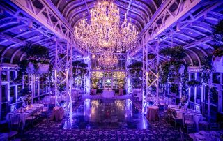 A grand, glass-ceilinged hall decorated with chandeliers, purple lighting, plants, and round dining tables arranged around a central dance floor.