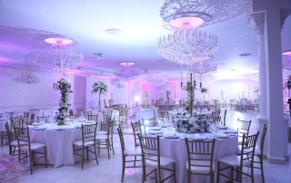 Elegant banquet hall set for an event with round tables, gold chairs, floral centerpieces, and crystal chandeliers illuminated by purple and white lighting.