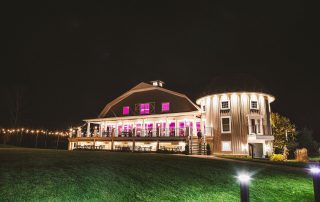 A large, well-lit barn-style building with purple lights in the windows stands on a grassy lawn at night.