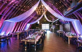A decorated barn venue with draped white fabric, string lights, chandeliers, and set tables arranged for an event or reception.